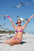 Woman wearing colorful bikini kneeling on beach with arms up, seagulls flying in the sky