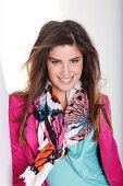 Pretty dark haired woman in pink jacket and colourful scarf leaning on wall, smiling