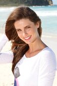 Portrait of cheerful dark haired woman wearing white top standing on beach, smiling