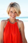 Portrait of pretty blonde woman wearing an orange top standing on beach, smiling