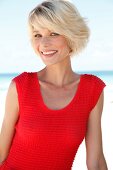 Portrait of pretty blonde woman with short hair wearing red top sitting on beach, smiling