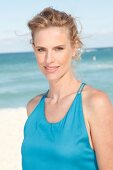 Portrait of beautiful blonde woman wearing turquoise top, standing on beach, smiling