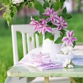 Vase mit Clematisblüten und Schälchen mit Apfelblüten auf Gartentisch