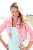 Portrait of beautiful blonde woman wearing short pink jacket standing on beach, smiling