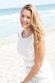 Portrait of happy blonde woman wearing white dress standing on beach, smiling