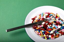 Different pills, tablets and capsules on plate with fork against green background