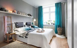 Schlafzimmer mit hellgrau getönten Wänden und blauen Vorhängen, langes Bücherbord über dem Doppelbett