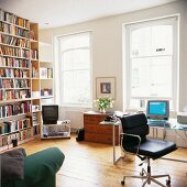 A wall of books across from a home office with a desk chair covered in black leather in front of a window in the living room