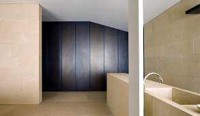 Monolithic stone counter with sink and free standing fitting in front of a wall covered with light colored stone tiles and dark vertical panels on the wall