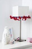 A white table lamp decorated with red feather ribbon and vases decorated with flowers