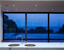 A shiny white marble surface with a kitchen sink in front of a window in a minimalistic room and a view of a pool by night