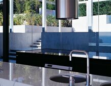 A sink set into a shiny stone work surface in a modern house with a view through a window onto a terrace