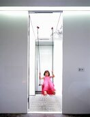 Blick durch Türöffnung auf Mädchen in pinkfarbenem Kleid, unscharf beim Schaukeln in lichtem Treppenraum
