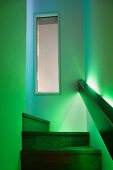 Durch kastenförmiges Geländer mit indirekter Beleuchtung in grünes Licht getauchter Treppenaufgang mit Holzstufen und Blick auf Innenfenster