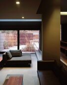 Offener Wohnraum in dunklen Tönen mit verschiedenen Sofas in tieferliegendem Sitzbereich und Blick durch Fensterfront in Garten