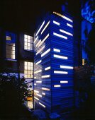 Tower extension with blue wood cladding and illuminated window strips in front of English house with brick facade