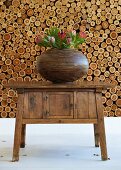 Brown striped, pot belly vase holding exotic protea flowers on old wooden wall table with neatly-stacked firewood in background