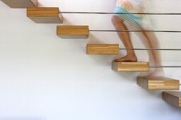 Blurred legs of bare-footed woman on floating staircase construction with wooden treads projecting from wall behind delicate baluster rods