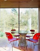 Rot gebeizte Holzstühle um Glastisch vor raumhohen Terrassenfenstern mit Ausblick