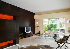 Modernes Wohnzimmer mit dunkler Holzwand und Tierfellteppich neben Couchtischen aus Acrylglas