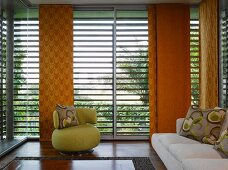 70s-style, small, green upholstered armchair with patterned cushion in front of louver blinds and orange sliding curtains