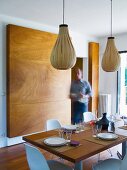 Man setting table in modern dining room with bulbous pendant lamps made from wooden strips