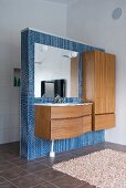 Minimalist vanity and wooden cupboard on a blue, mosaic tile dividing wall in a bathroom