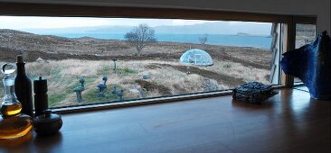 Blick aus querliegendem Küchenfenster auf karge Küstenlandschaft mit Skulpturen und Gewächshauskuppel