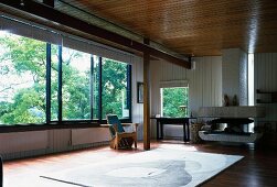 Open-plan living space in modern wooden house with wall and ceiling panels, large sliding windows and sculptural fireplace