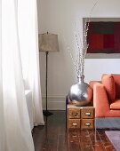 Silver-coloured vase on low, wooden cabinet and standard lamp in corner of living room
