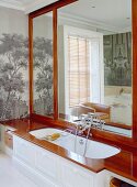Bathtub with wooden surround in front of large mirror & pictorial wallpaper