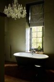 Dim period building bathroom with nostalgic chandelier and free-standing vintage bathtub below window