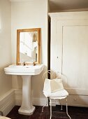 Delicate metal chair painted white next to antique pedestal washstand in corner of bathroom
