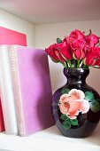 Strauss roter Rosen in altmodischer Keramikvase mit Rosenmotiv neben Büchern im Regalfach