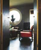 View through door into interior in dark shades with red armchair and sculpture in round window