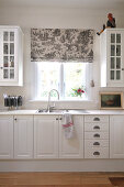 Simple, country-house-style kitchen unit with wall-mounted cupboards and sink below window with grey, patterned Roman blind