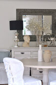 Loose covered chair at round dining table; lamp with silver base and male bust decorating vintage chest of drawers in background