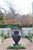 Fountain made from anthracite-coloured amphora on gravel garden path and view of road beyond white wooden fence