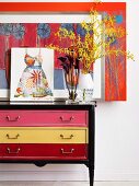 Vase of flowers and pictures on vintage chest of drawers with drawers of different colours against wall