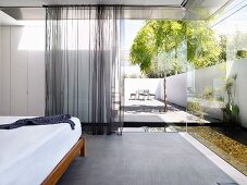 Designer bedroom with airy curtains at partially glazed facade with view of pool in sunny courtyard