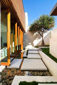 White stone slabs leading from living room across shallow pebble-filled pond to manicured garden