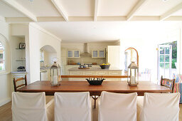 Langer Holztisch und Stühle mit Hussen, im Hintergrund offene Küche