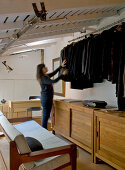 Schlafgalerie im Industriestil - Frau beim Suchen eines Kleidungsstücks an Stange mit dunklen Jacken, darunter Sideboard und Holzsofa mit hellen Polstern