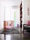 Helles, luftiges Babyzimmer mit Babybett auf weichem Wollteppich und fröhlichem, grünen Baum an der Wand