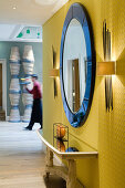 Round, wall-mounted mirror with blue frame between wall lamps on yellow, illuminated wall in open foyer with member of staff in background