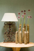 Group of ceramic vases holding single flowers and table lamp with white lampshade and vintage metal base on modern table against fabric-covered wall