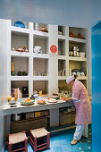 View through doorway of woman preparing food on kitchen counter in front of masonry shelving