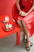 A woman wearing a red party dress sitting a small table with prawn sandwiches and a cocktail