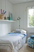 Einzelbett mit Tagesdecke in Zimmerecke neben Fenster und Ablage an weisser Holzwand