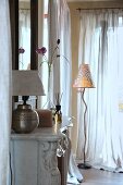 Table lamp on mantelpiece of grand open fireplace and modern standard lamp in corner of living room next to windows with airy curtains